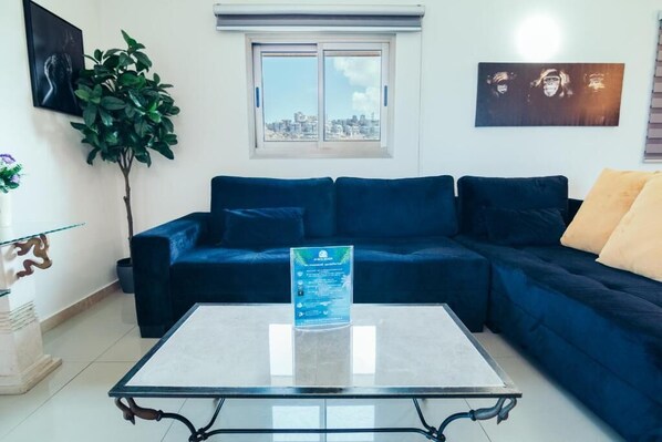 Un salon avec un canapé bleu foncé, une table basse en verre, une plante en pot et une vue sur la ville par la fenêtre.