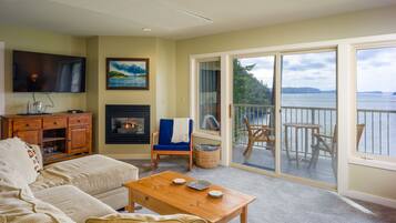 A living room with a sofa, a coffee table, a TV, a fireplace, and a balcony with a view of the water.