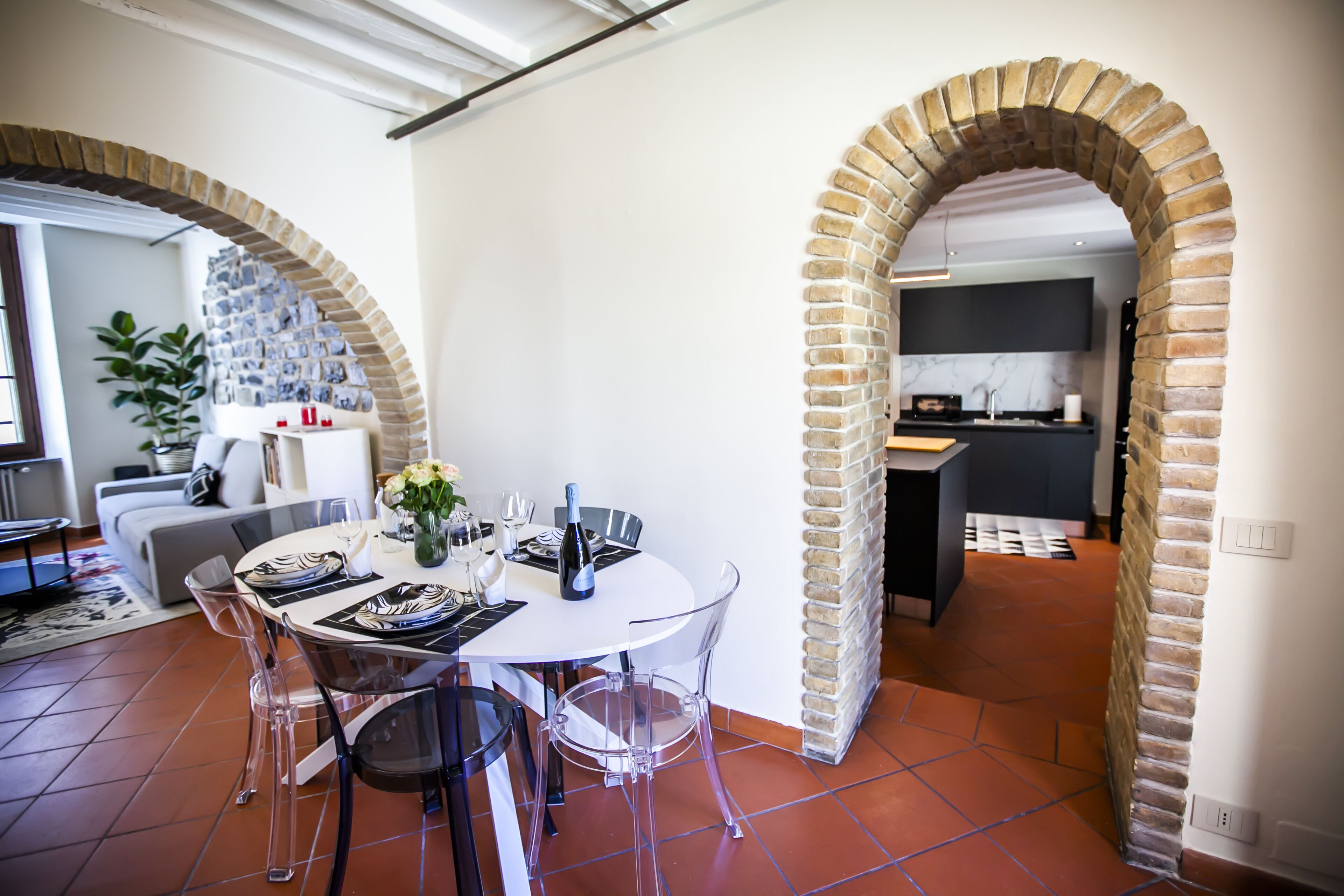 A dining area with a round table, clear chairs, and a brick archway leading to a kitchen.