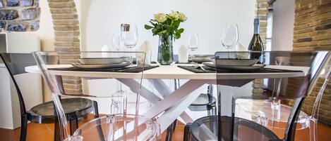 Uma mesa de jantar posta com copos e pratos de vidro, rodeada por cadeiras, em um ambiente com arcos de tijolos e piso ladrilhado.