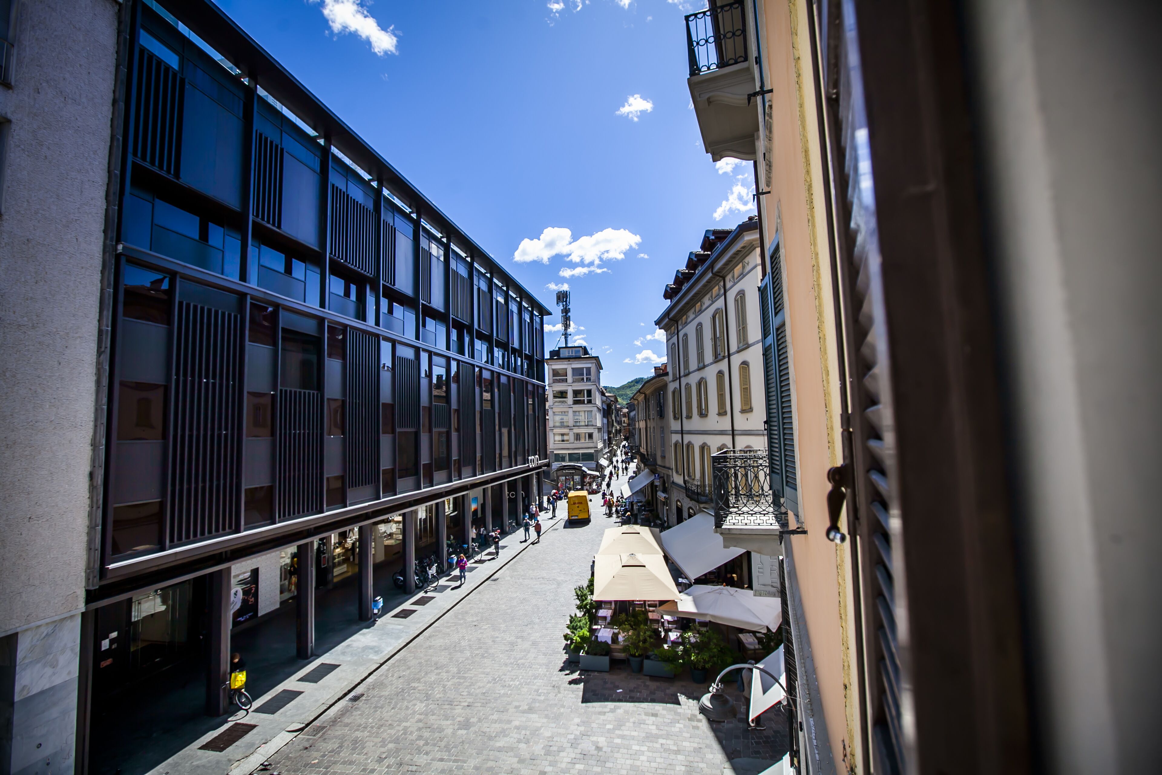 A narrow street with modern buildings, a yellow bus, and outdoor seating areas.