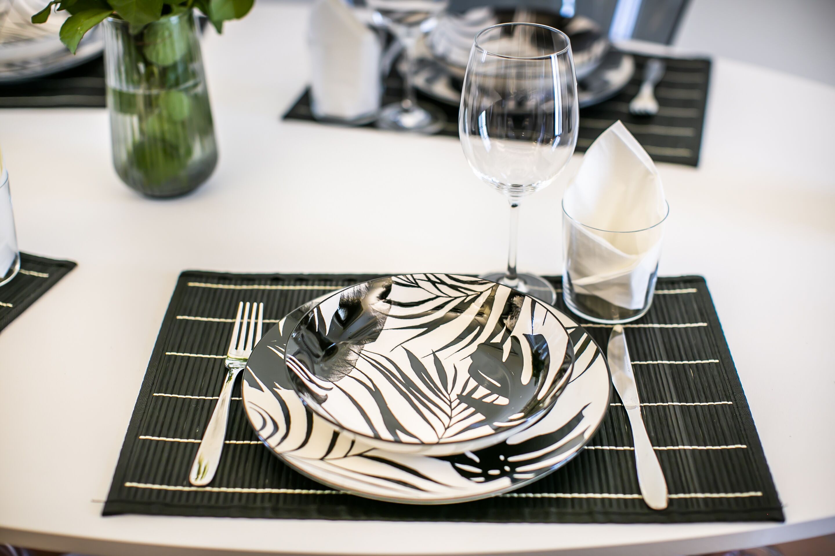Una mesa dispuesta con una servilleta, un plato blanco, un cuchillo, un tenedor y una copa de vino, todo sobre una base con estampado blanco y negro.