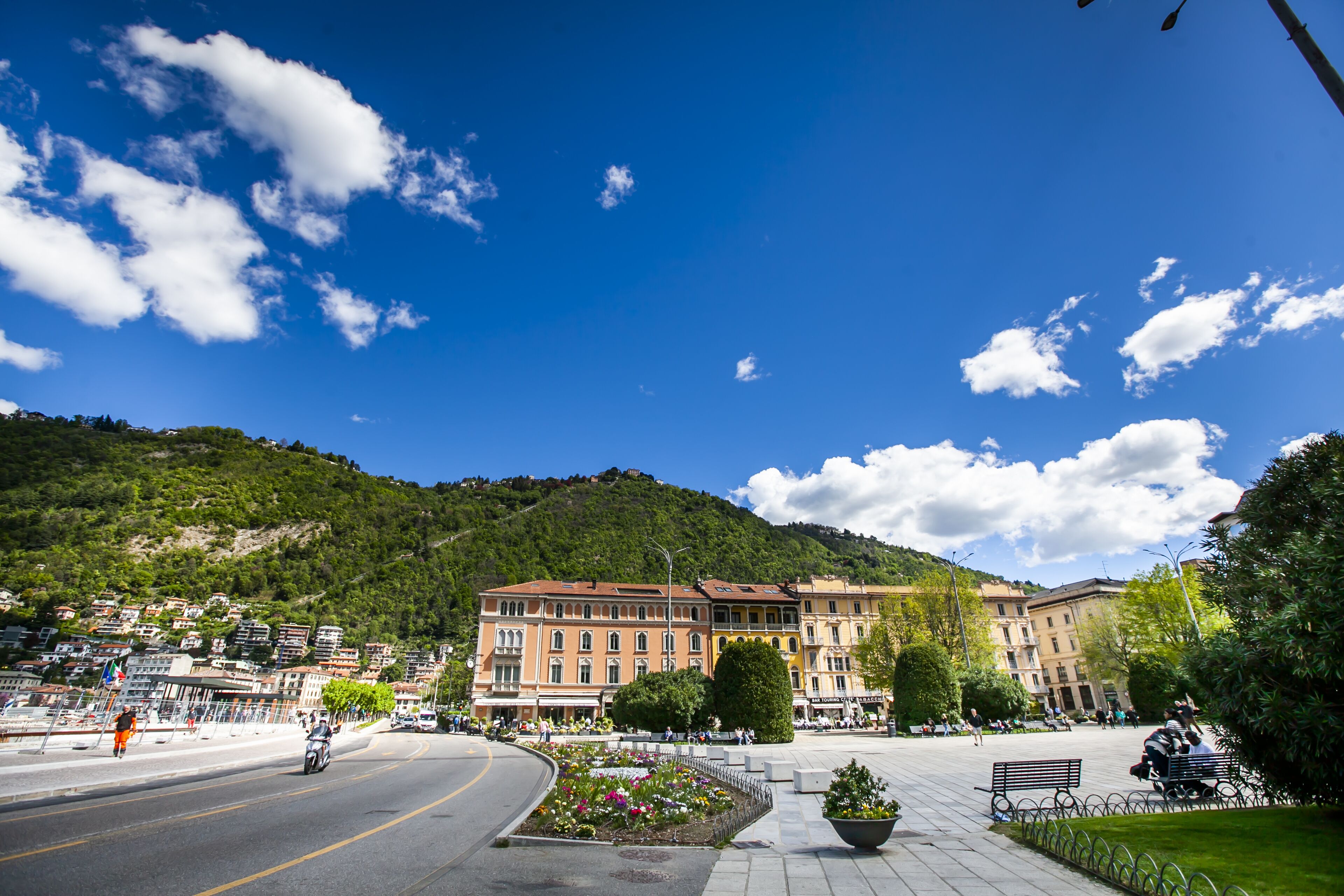 A town square with a large building, a flower garden, and a road with a motorcyclist.