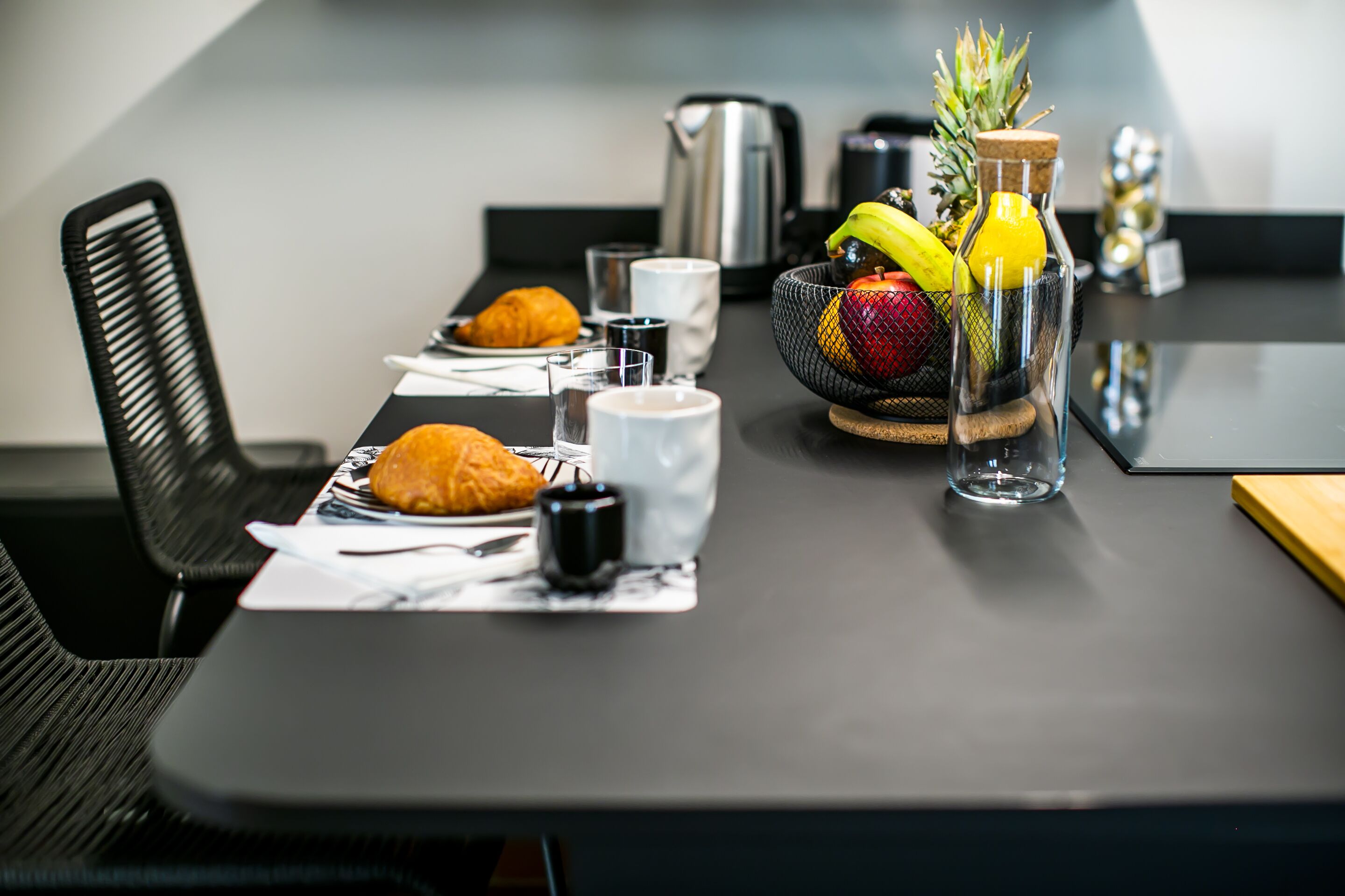 A breakfast setting with croissants, a fruit bowl, coffee cups, and a kettle on a modern table.