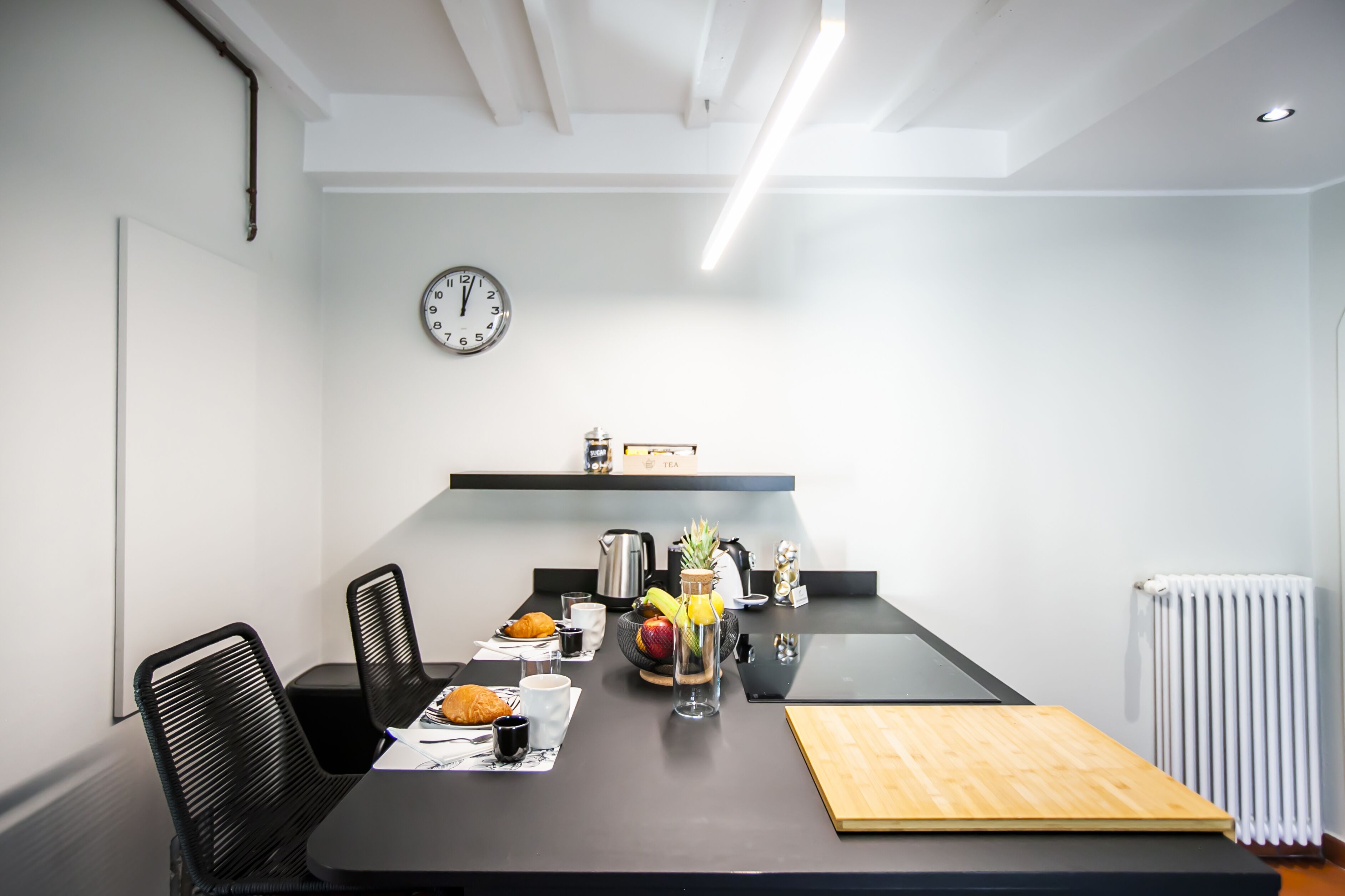A modern kitchen with a black countertop, white walls, and a clock. There are chairs, a table with food, and a radiator.