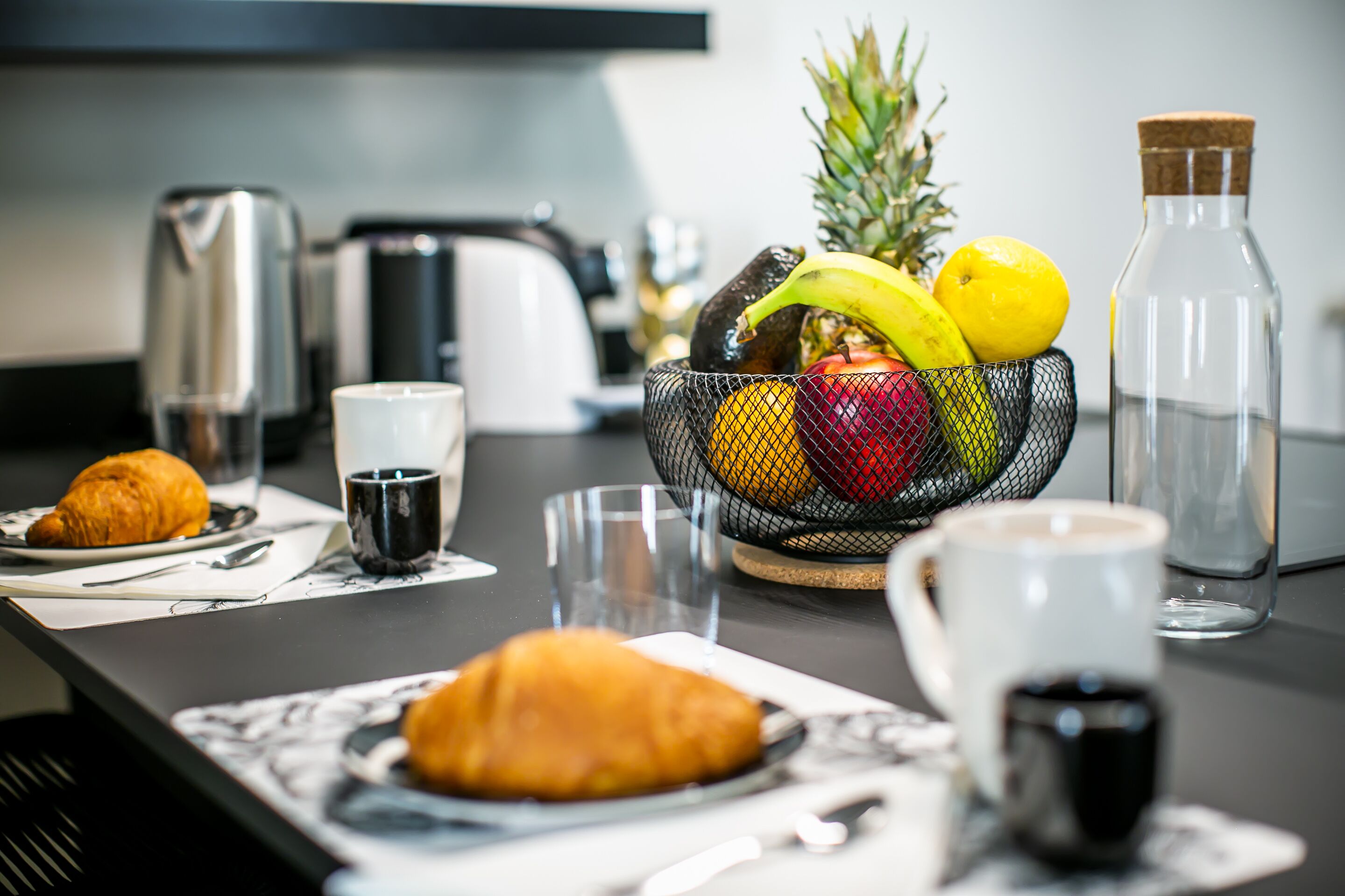 Una mesa con cruasanes, un frutero, tazas de café y una botella de agua.