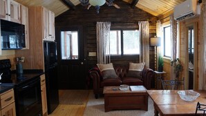 A cozy cabin-style room with wooden walls, a kitchenette, a leather sofa, a dining table, and a chandelier.