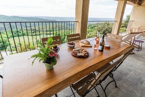 A wooden dining table with chairs, a bowl of fruit, a loaf of bread, and a bottle of wine on a balcony with a scenic view.