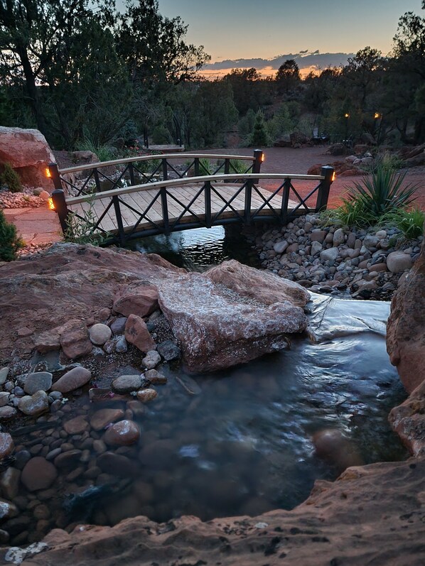 A wooden bridge over a stream with rocks and pebbles, surrounded by trees and a clear sky.