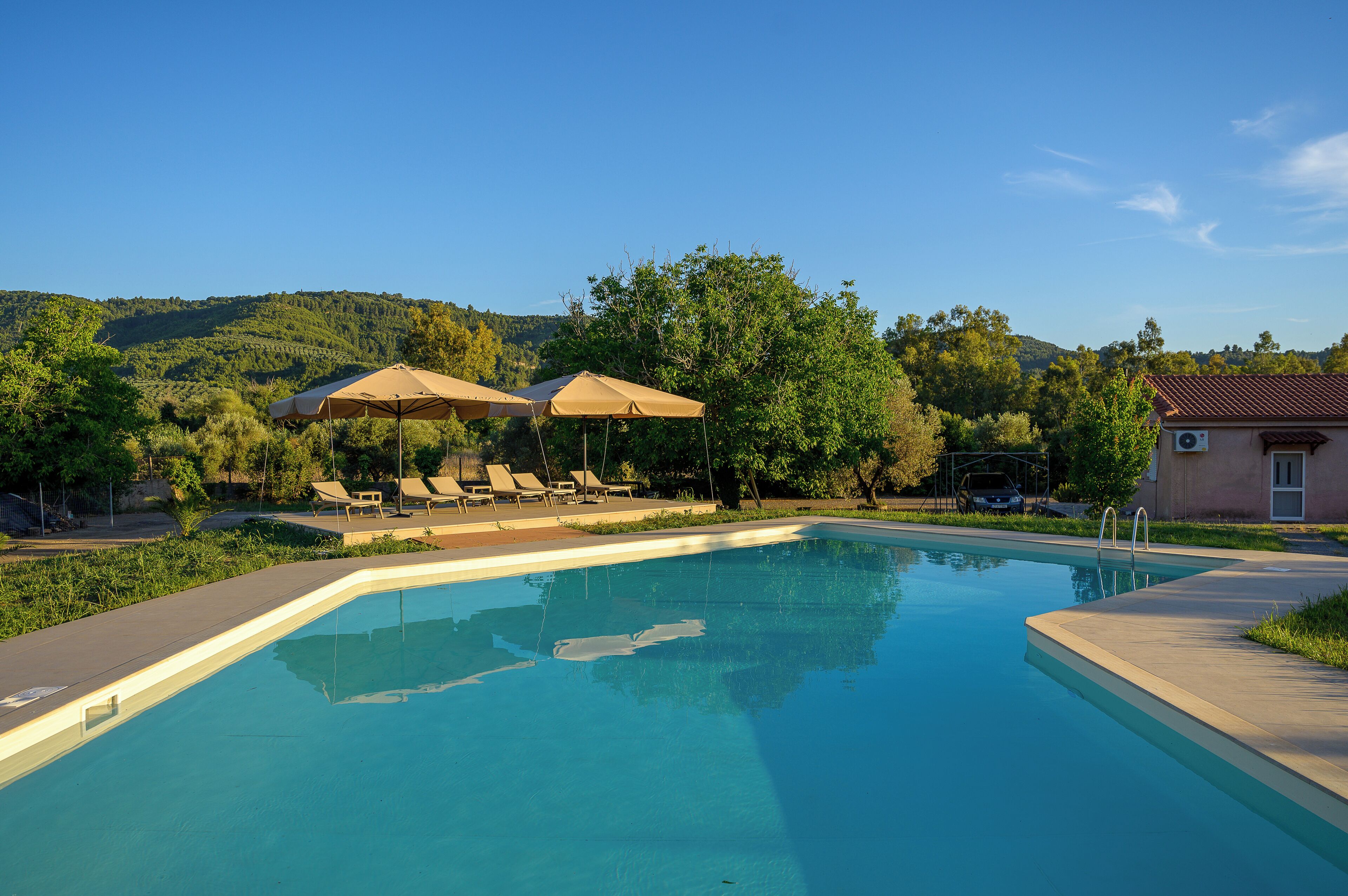 Una piscina con sillas de descanso y sombrillas, rodeada de vegetación y colinas.