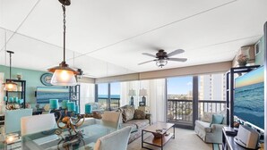 Ein geräumiges Wohnzimmer mit einem Glastisch, bequemen Sitzgelegenheiten, einem Deckenventilator und einem großen Fenster mit Blick auf den Ozean.