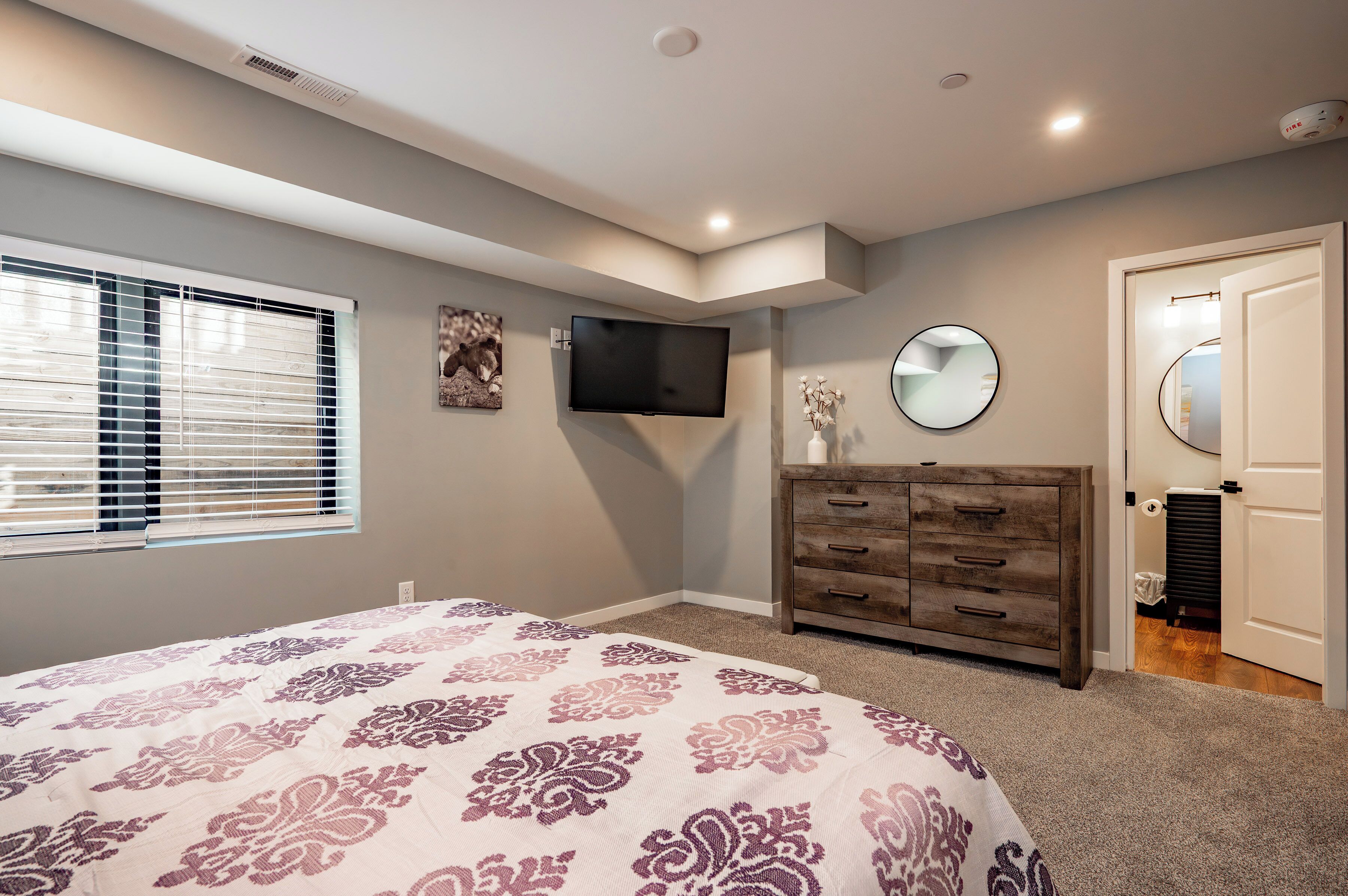 A bedroom with a bed, a dresser, a TV, a window with blinds, and a mirror.