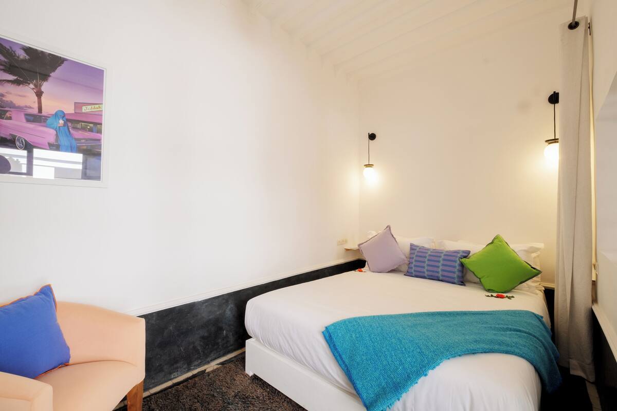 A bedroom with a bed, pillows, a blue blanket, a chair, and a wall-mounted light fixture.