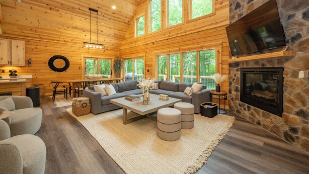 A log cabin interior with a stone fireplace, large windows, a sofa set, and a dining area. - New! Chic Chalet w/ Outdoor Fireplace & Hot Tub (McHenry)