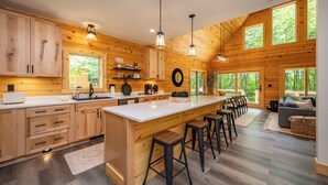 A spacious log cabin kitchen with a large island, bar stools, and a dining area with a view of the forest. - New! Chic Chalet w/ Outdoor Fireplace & Hot Tub (McHenry)