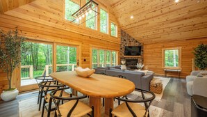 A wooden cabin interior with a dining table, chairs, a gray sofa, and a stone fireplace. - New! Chic Chalet w/ Outdoor Fireplace & Hot Tub (McHenry)