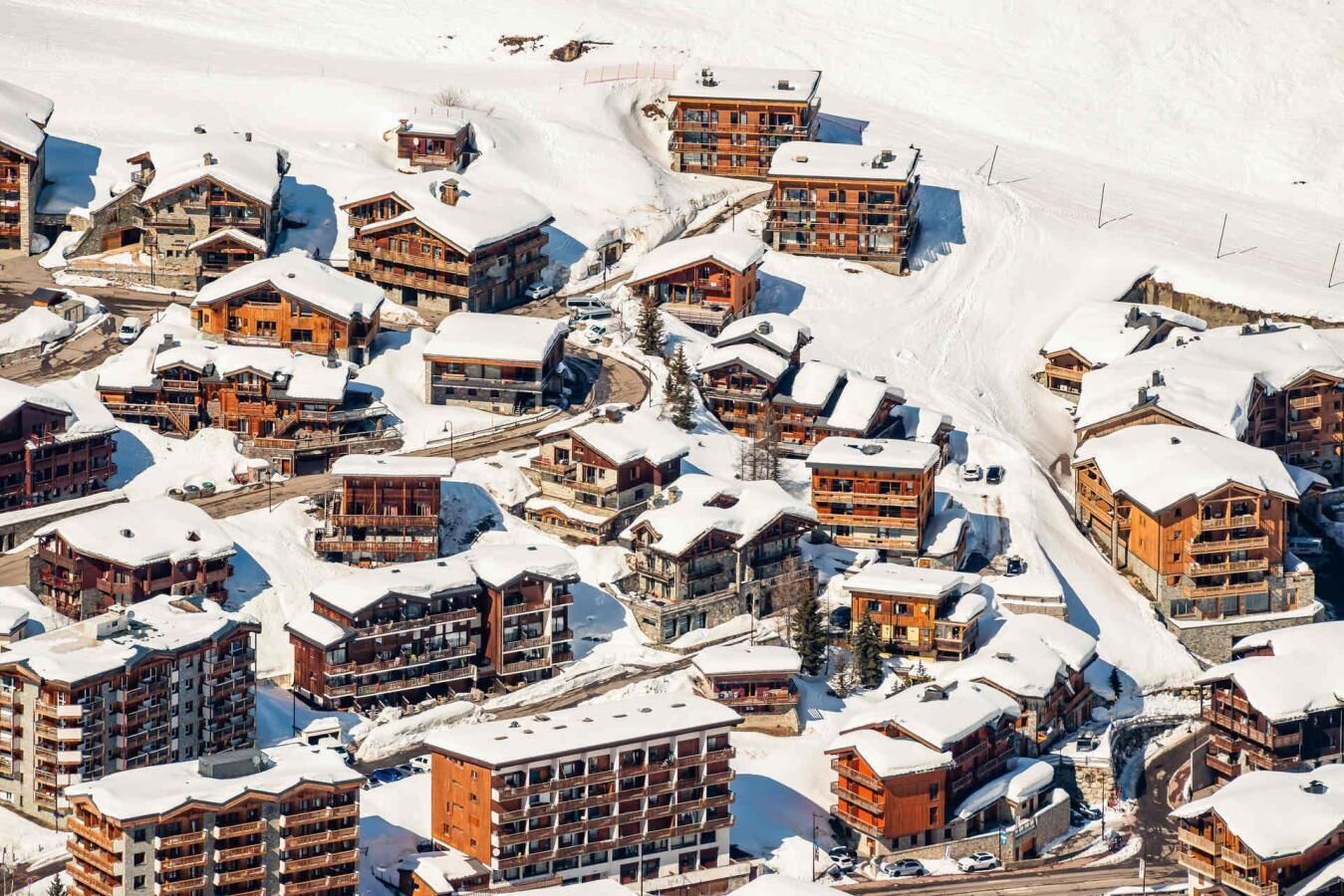 Estância de esqui com vista para uma montanha coberta de neve, com vários edifícios em estilo chalé, teleféricos e uma estrada sinuosa.