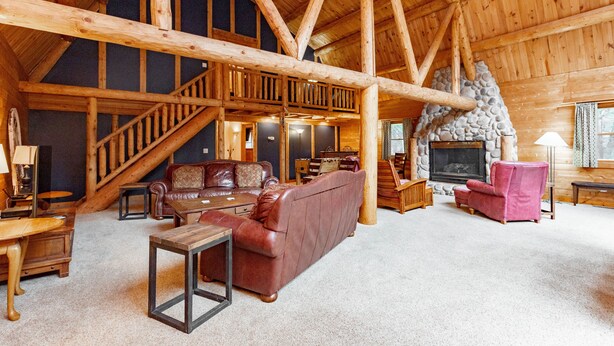 A spacious loft-style living area with a high ceiling, wooden beams, a fireplace, and a staircase leading to an upper level. - Dog Friendly Lodge w/ Hot Tub & Views (McHenry)
