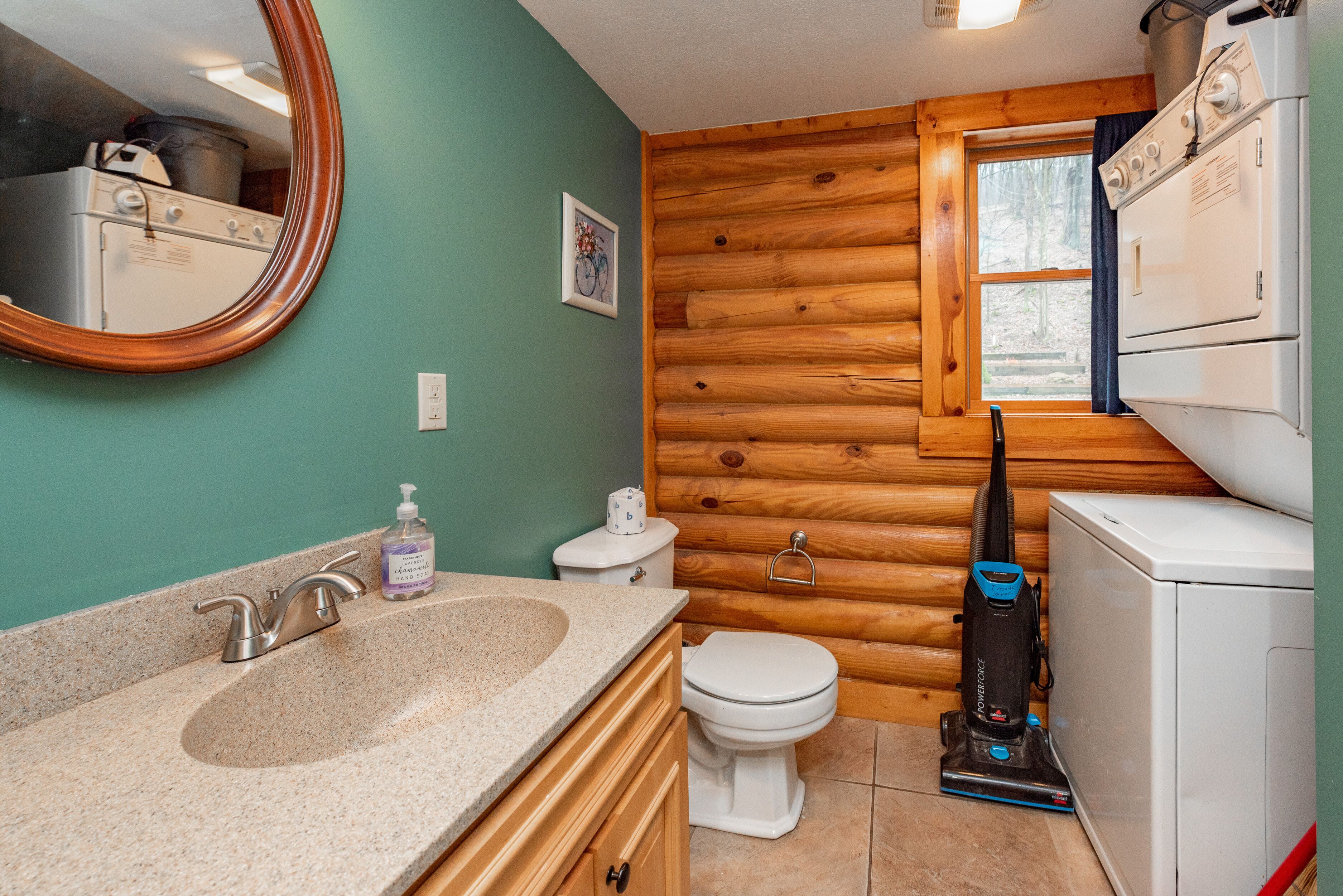 A bathroom with a wooden log wall, a sink with a mirror, a toilet, and a vacuum cleaner.