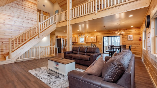 A spacious living area with a wooden staircase, leather sofas, a dining table, and a kitchen in the background.