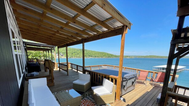 A covered deck with a wooden floor, white cushioned seating, and a table. - AMAZING WATERFRONT HOME WITH HOT TUB. BOTH HAVE EXTENSIVE VIEWS OF THE LAKE (Cookson)