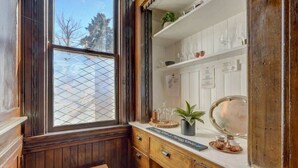 Ein Zimmer mit Holzwänden, einem Fenster, einem kleinen Hocker, einer weißen Arbeitsplatte mit Glaswaren und einem Spiegel.
