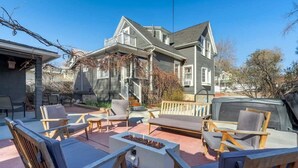 Ein zweistöckiges Haus mit überdachter Terrasse, auf der Gartenmöbel und eine Feuerstelle Platz finden.