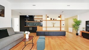 A modern living room with a grey sofa, a small table with a vase, and a blue ottoman. There is a kitchen area with black cabinets and a tiled backsplash in the background. - Superb Appartement: 3br/8p - Père Lachaise (Paris)