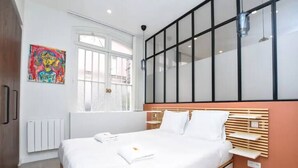 Une chambre moderne avec un grand lit, une fenêtre avec des stores, un radiateur et un tableau coloré accroché au mur.