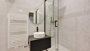 A modern bathroom with a glass-enclosed shower, a round mirror, and a white sink on a black countertop. - Charming Appart 4p/1br G Le Marais (Paris)