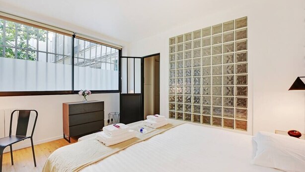 A modern bedroom with a large bed, a desk, and a view through a glass block wall. - Charming Appart 4p/1br G Le Marais (Paris)