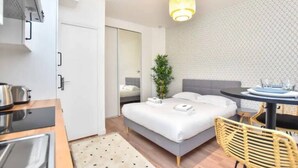 A compact hotel room with a bed, a dining table, a small kitchen area, and a plant.