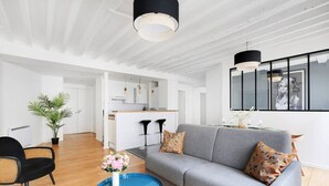A modern living room with a grey sofa, a round blue coffee table, and a black chair. There is a kitchen area in the background with a white countertop and black bar stools.