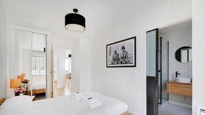 A modern hotel room with a large bed, a glass shower enclosure, a framed photograph on the wall, and a wooden bedside table with a lamp.