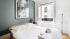 A neatly made bed with white linens, a framed photograph on the wall, a bedside lamp, and a view of a balcony through an open window.