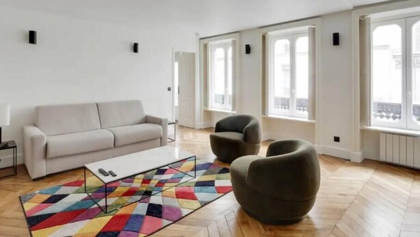 Een ruime woonkamer met een witte bank, een salontafel en twee fauteuils. Grote ramen zorgen voor natuurlijk licht en uitzicht.