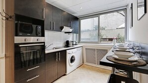 Une cuisine moderne avec des meubles de couleur sombre, un four encastré, un four à micro-ondes et un lave-linge. Une grande fenêtre offre une lumière naturelle et une vue sur l’extérieur.
