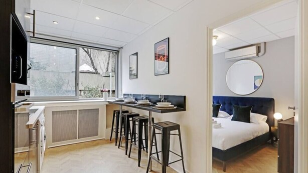 Une chambre d’hôtel compacte avec une kitchenette, un lit doté d’une tête de lit bleue, un miroir rond et une fenêtre donnant sur un paysage agréable.