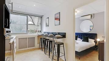A compact hotel room with a kitchenette, a bed with a blue headboard, a round mirror, and a window with a view.