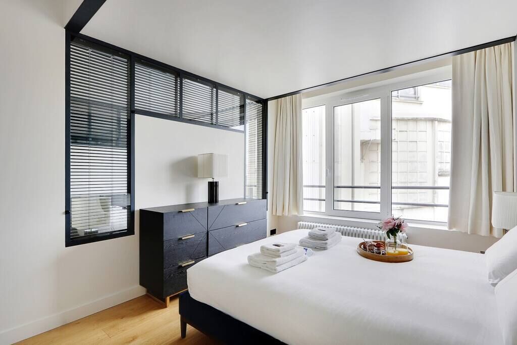 A modern bedroom with a large bed, a dark dresser, a bedside table, and a window with curtains.