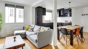 Um apartamento moderno com sala de estar, um sofá cinzento, uma mesa de jantar de madeira com cadeiras e cozinha com armários escuros e luminária pendente.
