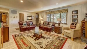 Una sala de estar acogedora con dos sofás de cuero, una mesa de centro y un sillón beige. Una alfombra grande con una escena de bosque cubre el suelo.