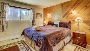 Un dormitorio con una cama grande, techo de madera, dos lámparas de noche, un ventanal con cortinas y una mesita de noche pequeña.