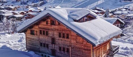 A wooden chalet with a balcony, surrounded by snow-covered trees and a ski slope in the background.