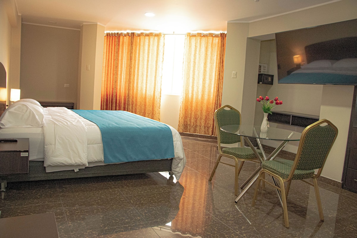 A hotel room with a bed, a glass table with chairs, a TV, and a window with curtains.
