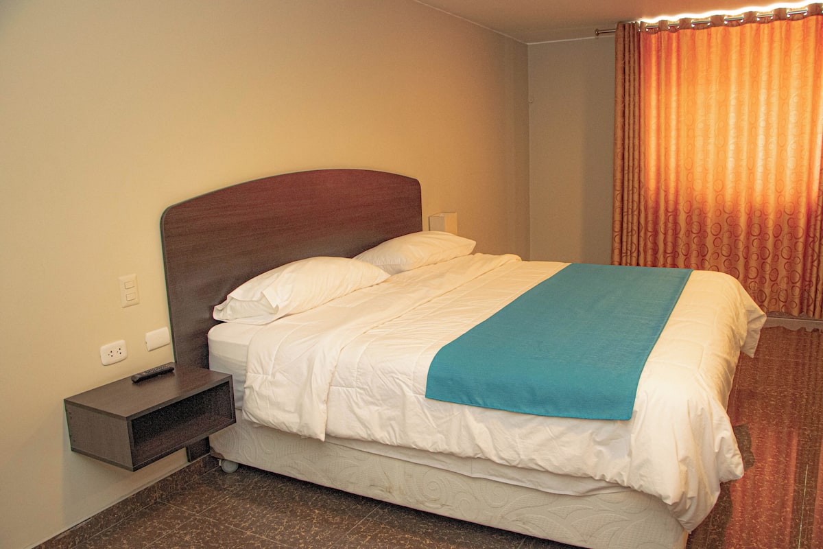 A neatly made bed with a wooden headboard, a bedside table with a remote control, and a curtain partially drawn to the side.