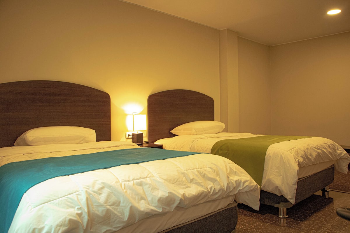 A hotel room with two beds, each with a headboard, a nightstand with a lamp, and a carpeted floor.