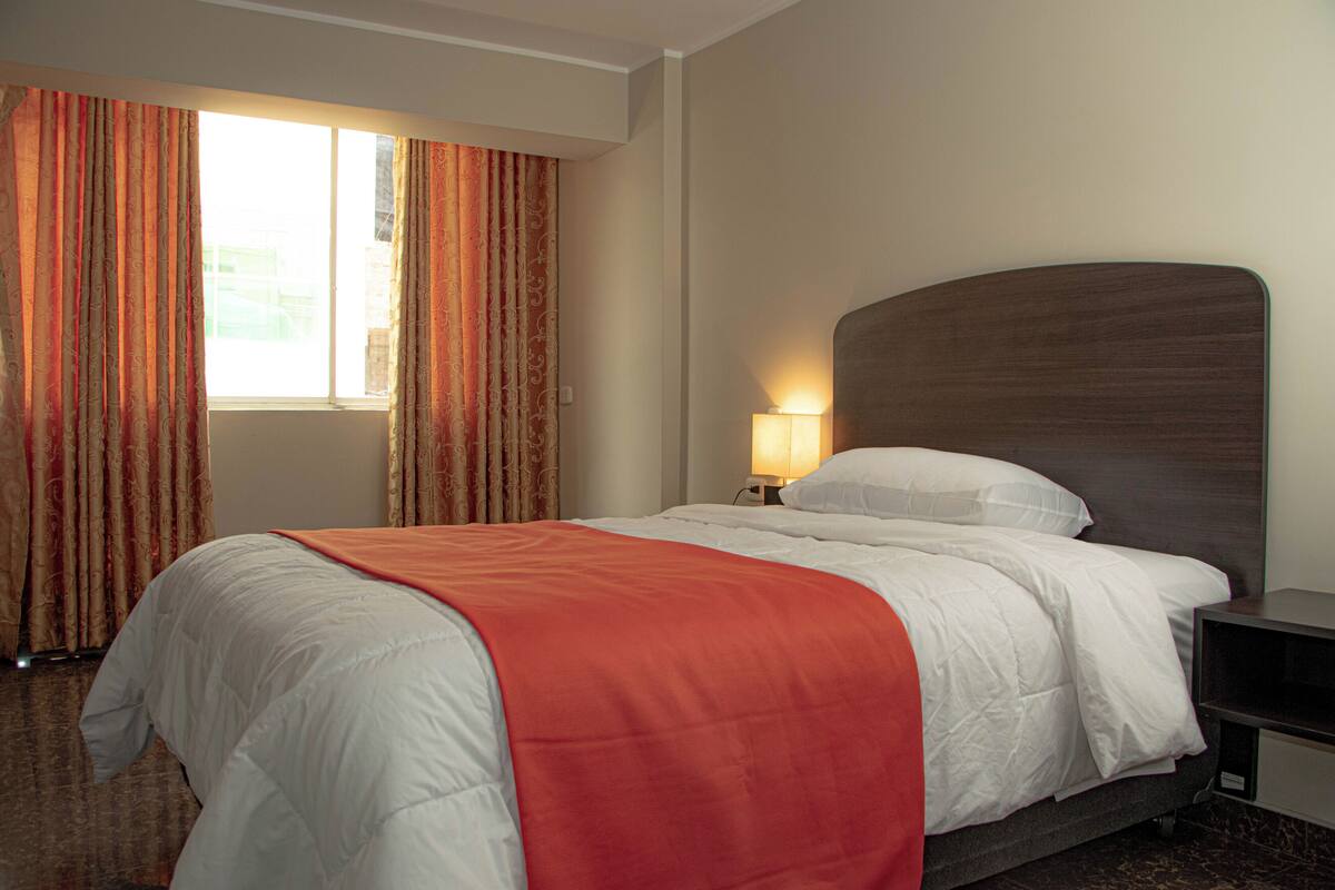 A hotel room with a large bed, a nightstand with a lamp, and a window with curtains.