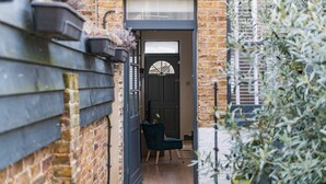 A narrow alleyway leading to a dark door with a glass panel, a green chair outside, and a brick wall with a window. - The Little Cottage, Whitstable (Whitstable)