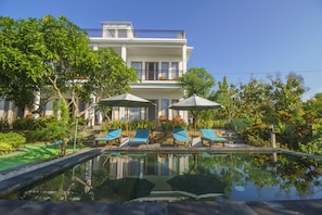 A pool with lounge chairs and umbrellas, surrounded by greenery and a building with a balcony. - Govardhan Bungalow (Penida Island)